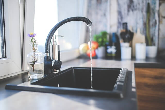 running faucet with a tasteful dribble of water pouring out of the tap in a well lit modern kitchen next to a single lavender flower with splashes of orange and green vegetables in the blurred background